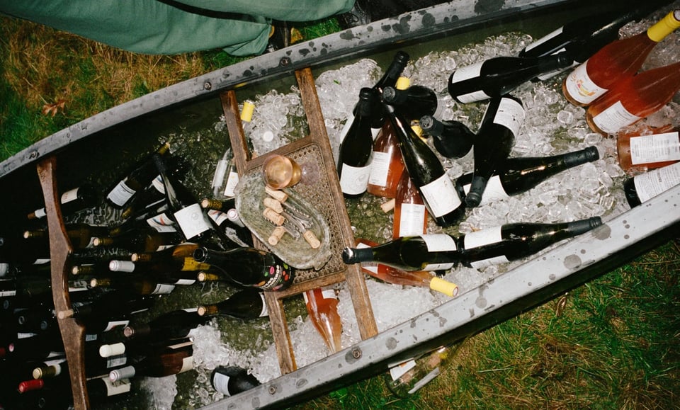 Around 50 various bottles of wine sitting in ice placed within a small metal boat sitting on the grass.