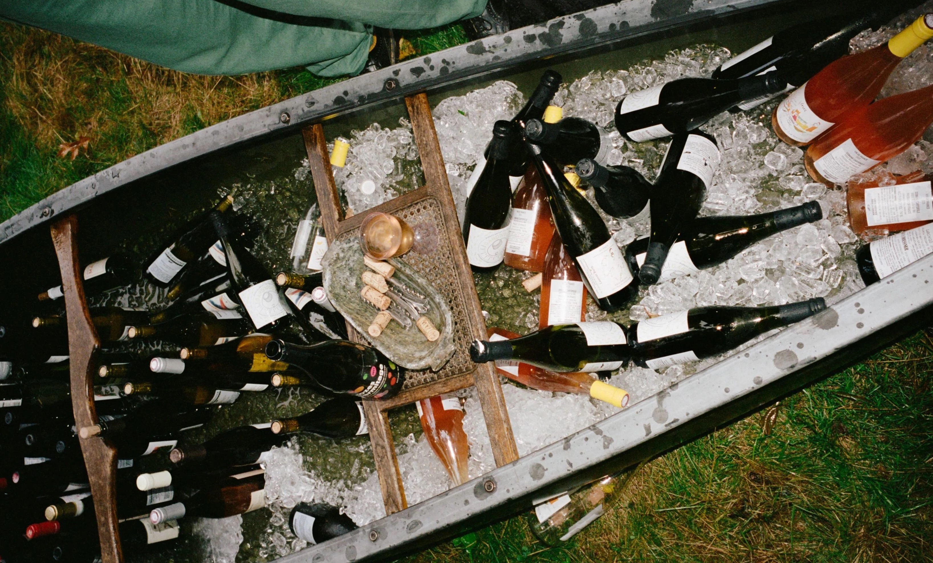 Around 50 various bottles of wine sitting in ice placed within a small metal boat sitting on the grass.